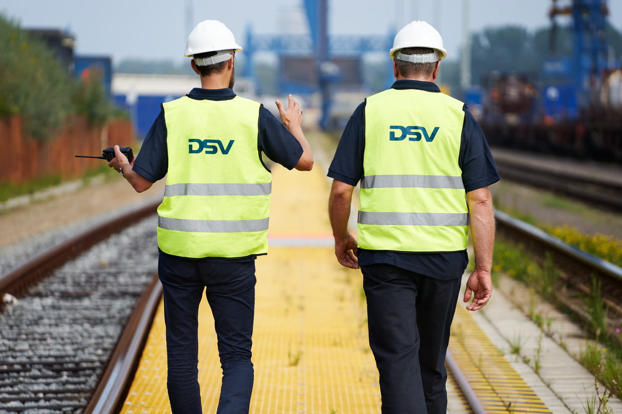 To DSV-medarbeidere ikledd refleksvest og vernehjelm går langs togskinne